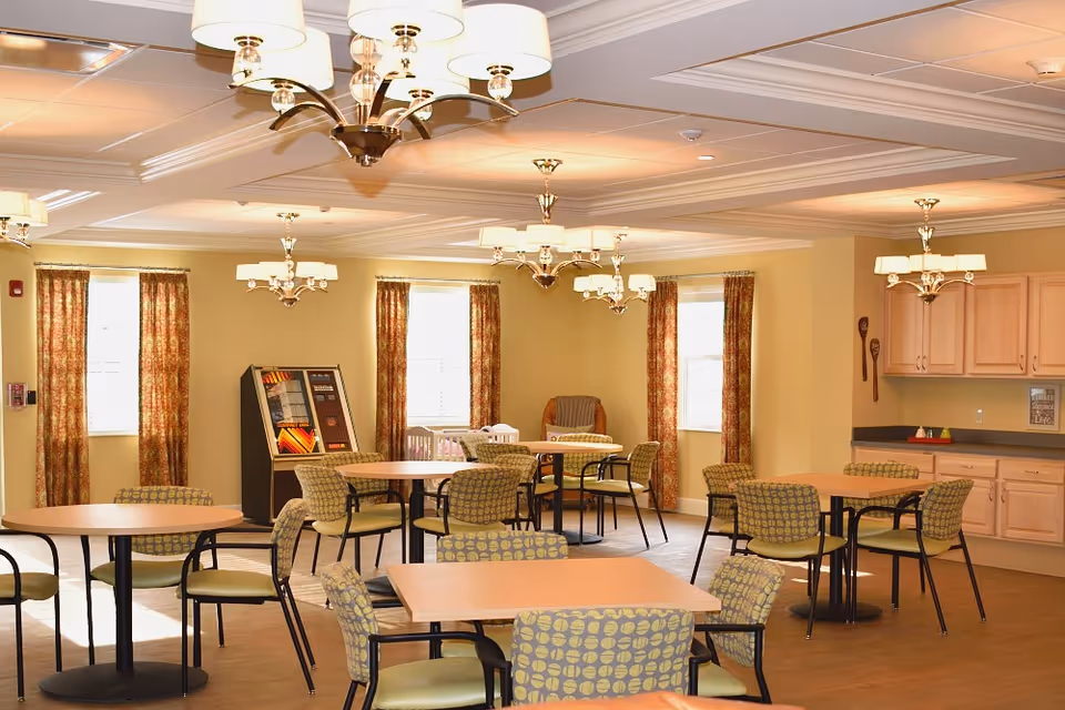 A well-lit dining room with multiple round and square tables surrounded by patterned chairs. The room features beige walls, several windows with floral curtains, and multiple ceiling light fixtures. There is a jukebox against one wall and a kitchenette area with light wood cabinets and a countertop.