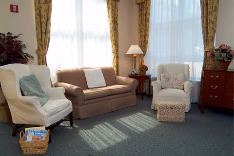 A cozy living room area with a beige sofa, two white armchairs, one with a green pillow and the other with floral cushions and a matching ottoman. There is a wooden side table with a lamp and a flower arrangement, floral curtains on the windows, and a basket with magazines on the floor.