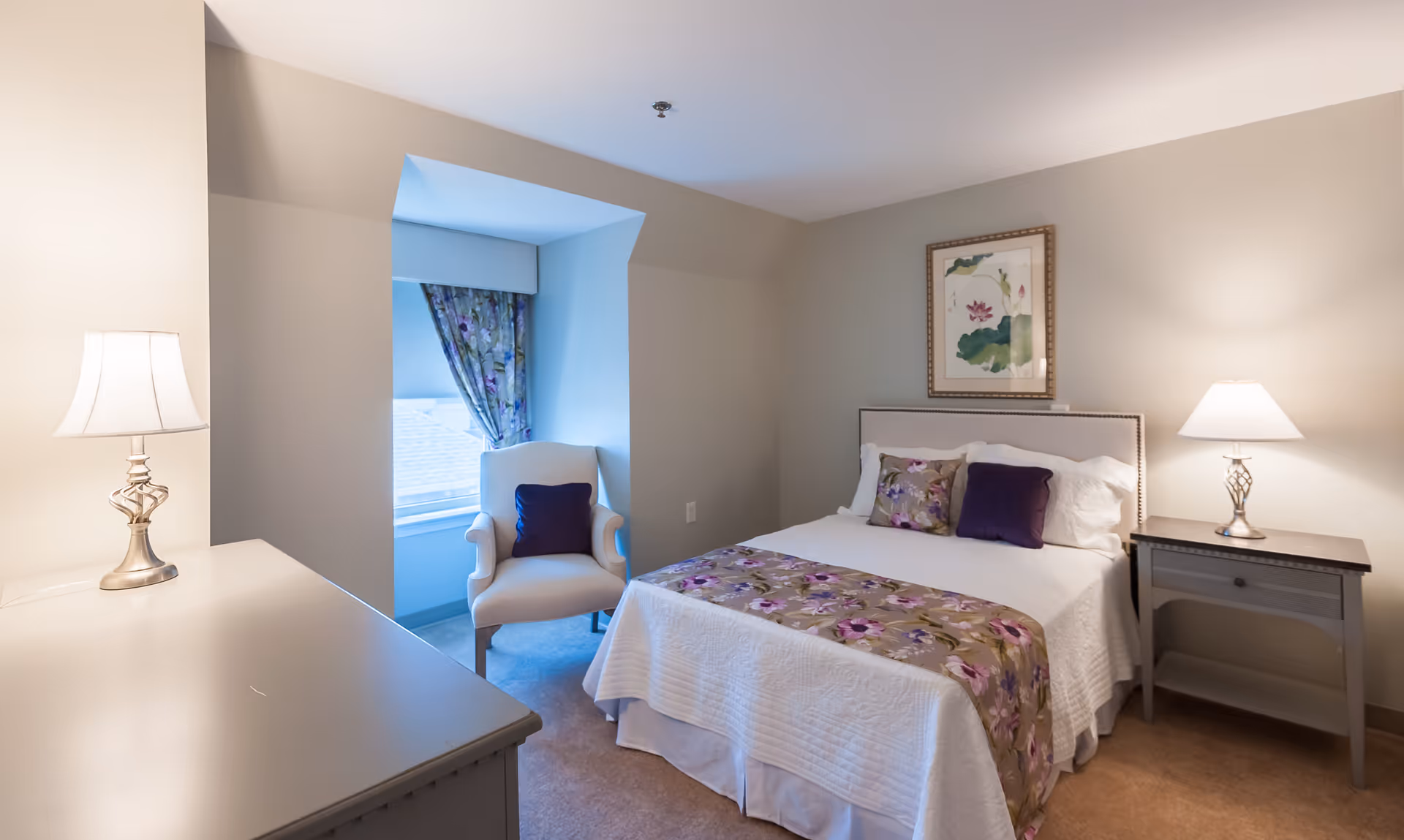 A cozy bedroom with a neatly made bed featuring white bedding and floral accents. There are two bedside tables, each with a lamp, and a comfortable armchair by a window with floral curtains. A framed artwork hangs above the bed.