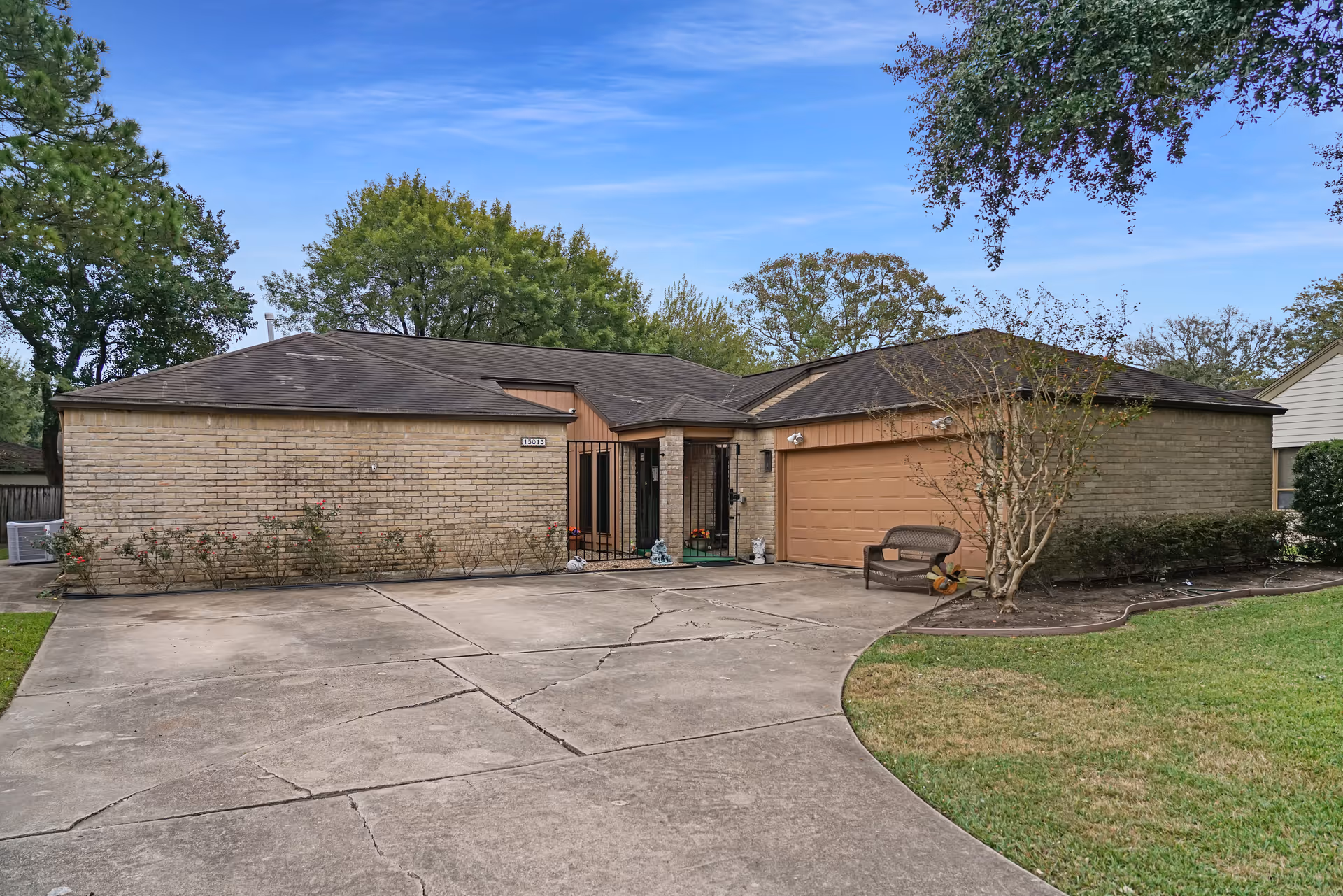 Exterior view of a single-story brick building with a two-car garage and a concrete driveway. There is a small bench and some landscaping with bushes and trees around the building under a blue sky.