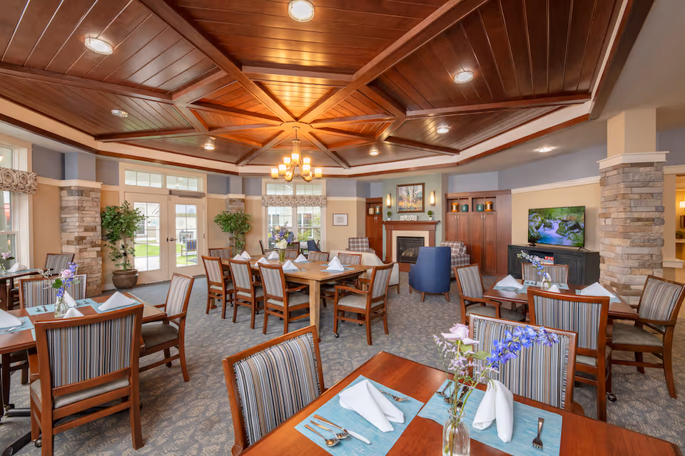 A spacious dining room with wooden tables and striped cushioned chairs neatly arranged. Each table is set with folded white napkins, silverware, and small floral centerpieces. The room features a decorative wooden ceiling with recessed lighting, stone pillars, large windows, and a fireplace with a painting above it. A television is mounted on a cabinet to the right, and there are potted plants near the windows and doors leading outside.