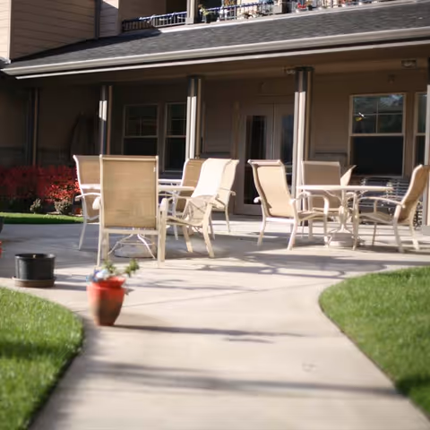 Outdoor patio area with several beige chairs and tables on a concrete surface, adjacent to a building with multiple windows and a door. There are green grass lawns on either side of the walkway leading to the patio, and a small red flower pot is visible in the foreground.