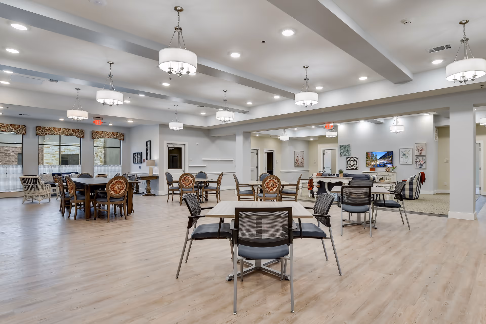 Spacious and well-lit common area in a senior living facility with multiple tables and chairs arranged for dining or social activities. The room features modern light fixtures hanging from the ceiling, large windows with decorative valances, and a seating area with a TV and artwork on the walls.