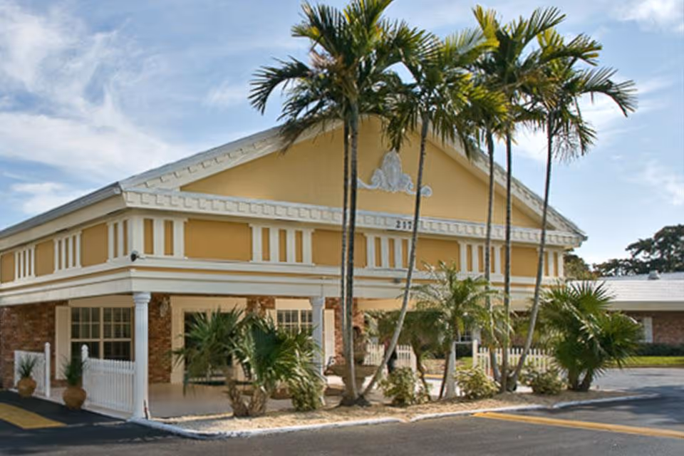 Exterior view of Darcy Hall of Life Care building with yellow and white facade, palm trees in front, and a paved parking area.