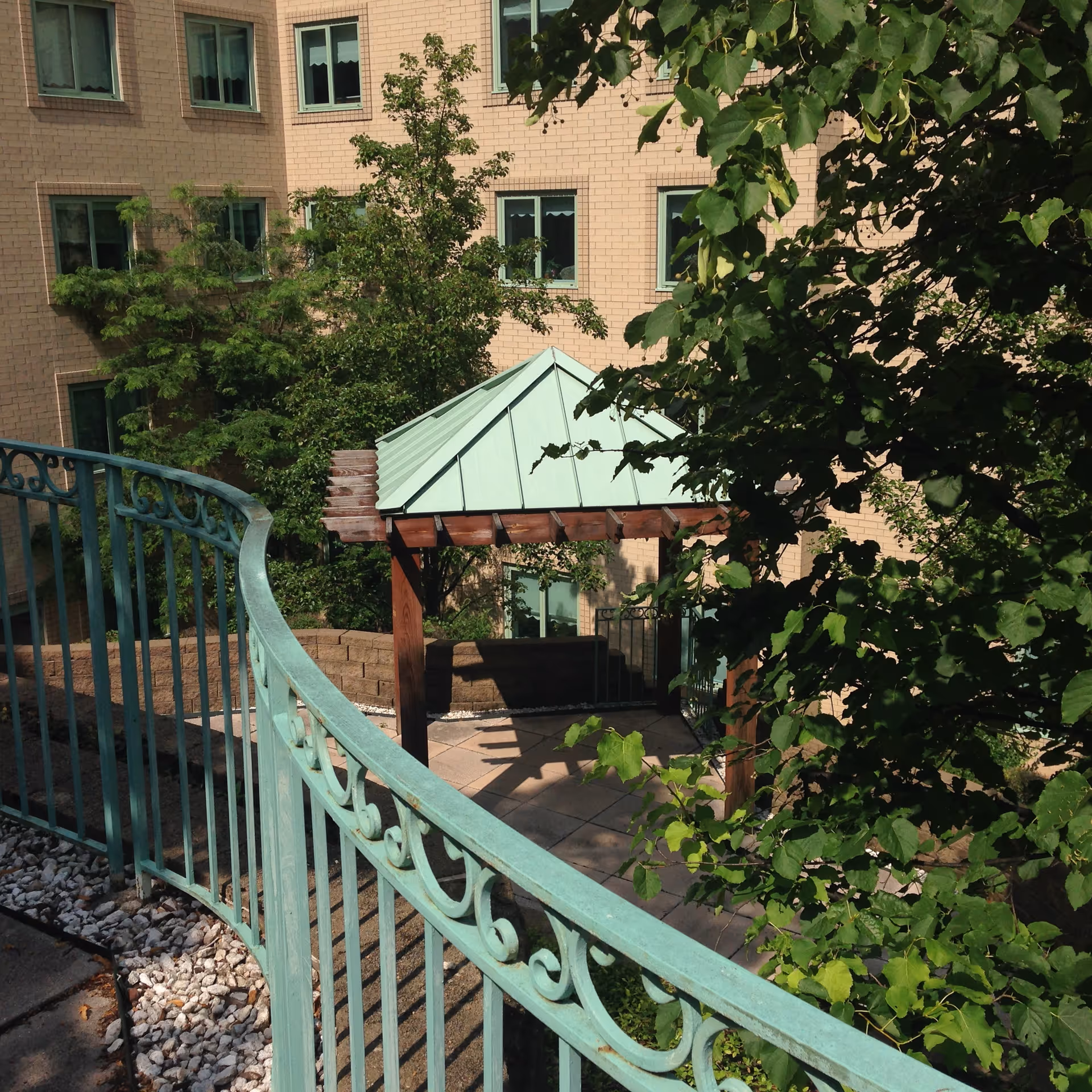 Outdoor area at Vincentian Schenley Gardens featuring a curved green metal railing, a small pavilion with a green roof supported by wooden beams, surrounded by trees and greenery, with a beige brick building in the background.