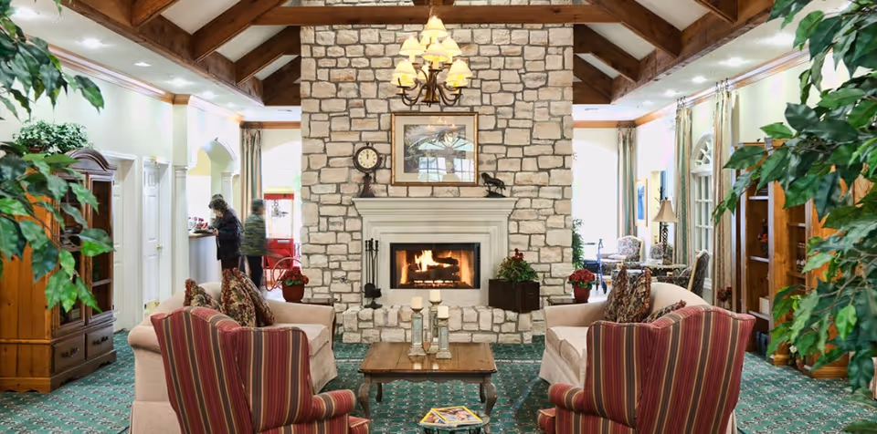 A cozy living room area in a senior living facility with a stone fireplace at the center, flanked by two beige sofas and two striped armchairs around a wooden coffee table. The room features a green patterned carpet, wooden ceiling beams, plants, and a chandelier above the fireplace. Two people are seen near the back, one standing and one walking.