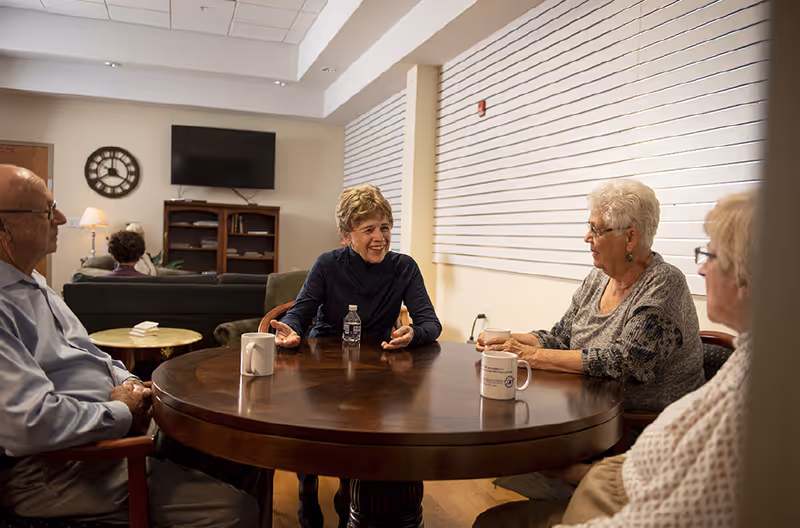 Four elderly individuals sitting around a round wooden table in a well-lit room, engaged in conversation. There are mugs and a water bottle on the table. In the background, there is a wall-mounted TV, a clock, a bookshelf, and a lamp beside a couch.
