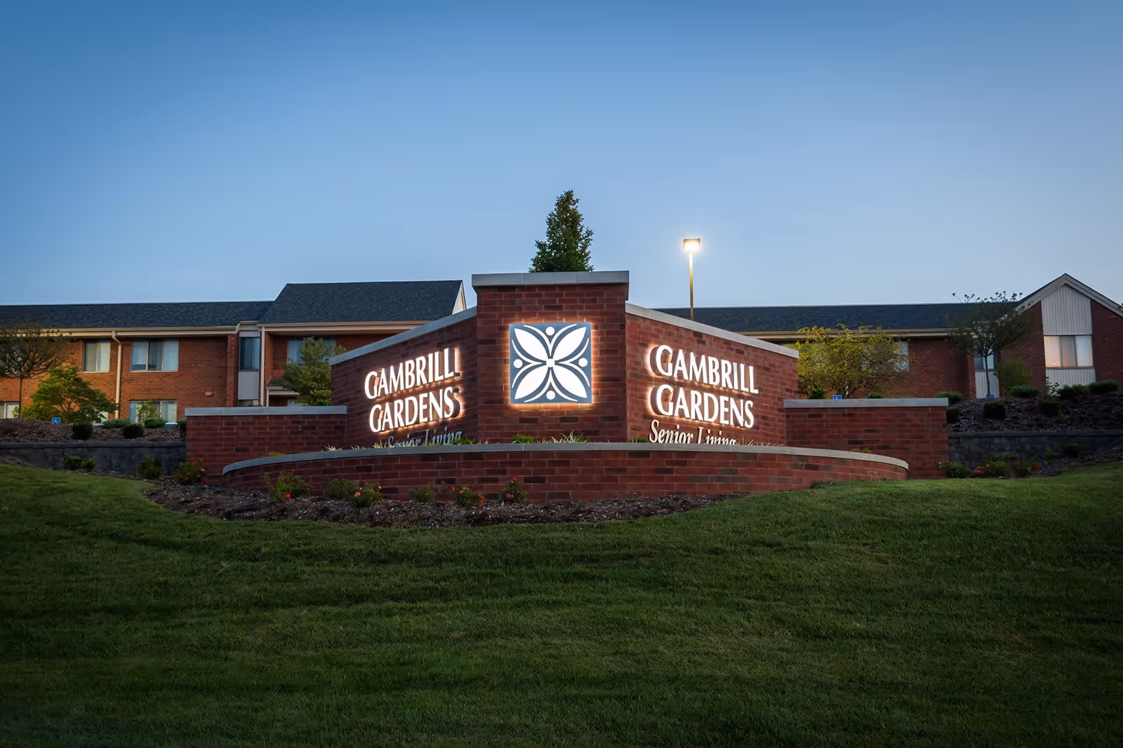 Illuminated brick sign for Gambrill Gardens Senior Apartments in front of a two-story brick building during dusk with a well-maintained lawn and some small trees and shrubs.