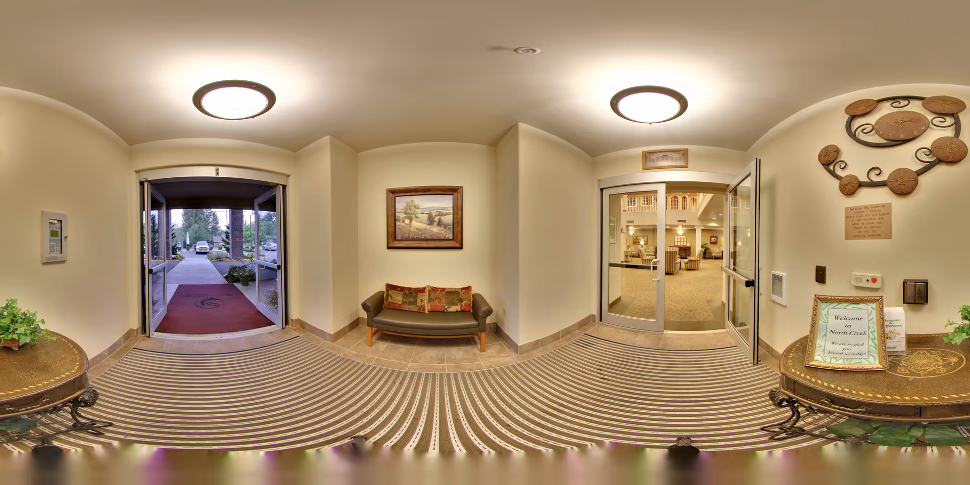 Entryway of a senior living facility with a glass door leading outside to a pathway and parking area. Inside, there is a small seating area with a bench and decorative pillows, a framed landscape painting on the wall, and two round tables with plants and a welcome sign for North Creek by Bonaventure. The interior beyond the glass door shows a lounge area with chairs and warm lighting.