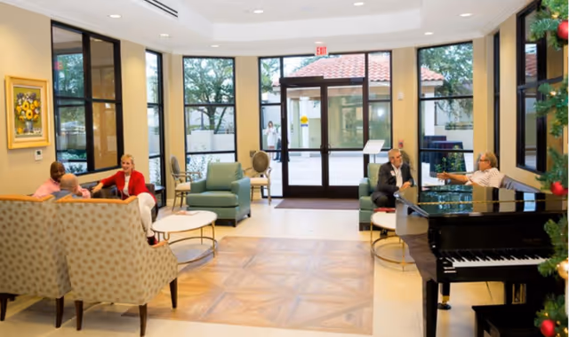 A bright and spacious living room area in a senior living facility with large windows and glass doors letting in natural light. The room features comfortable seating including armchairs and sofas arranged around small coffee tables. A black grand piano is positioned on the right side near a decorated Christmas tree. Four people are seated and engaged in conversation. Outside, trees and a tiled roof are visible through the glass doors.
