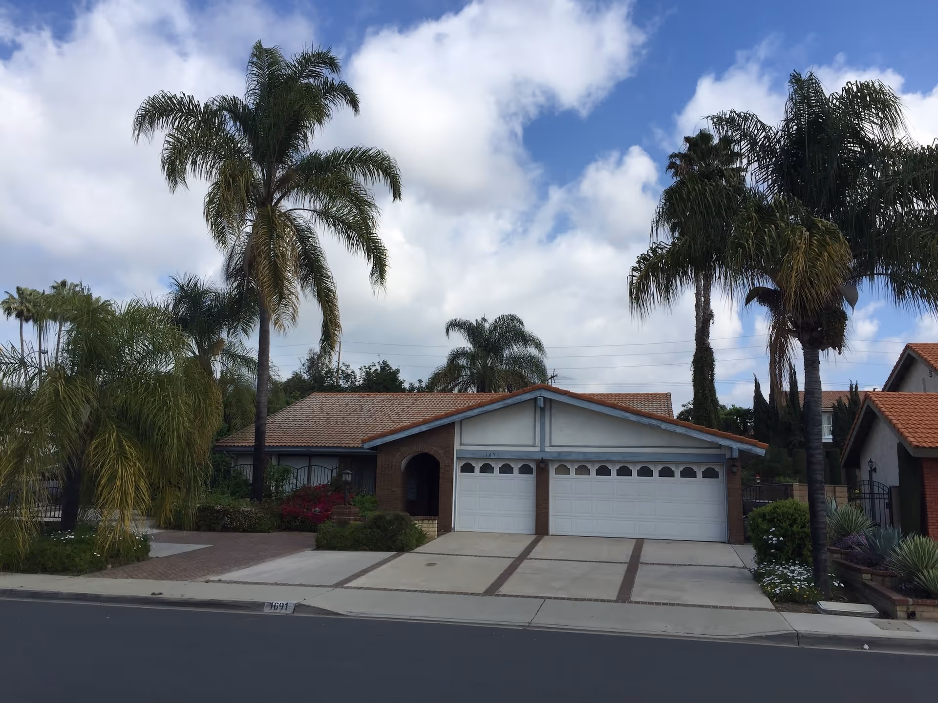 Single-story residential house with a three-car garage, surrounded by tall palm trees and other greenery under a partly cloudy sky.