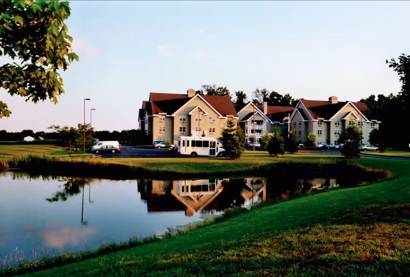 View of a senior living facility named Parkrose Estates with multiple beige buildings featuring peaked roofs, surrounded by green lawns and trees. A small white shuttle bus and several cars are parked near the buildings. In the foreground, there is a pond reflecting the buildings and trees under a clear sky.