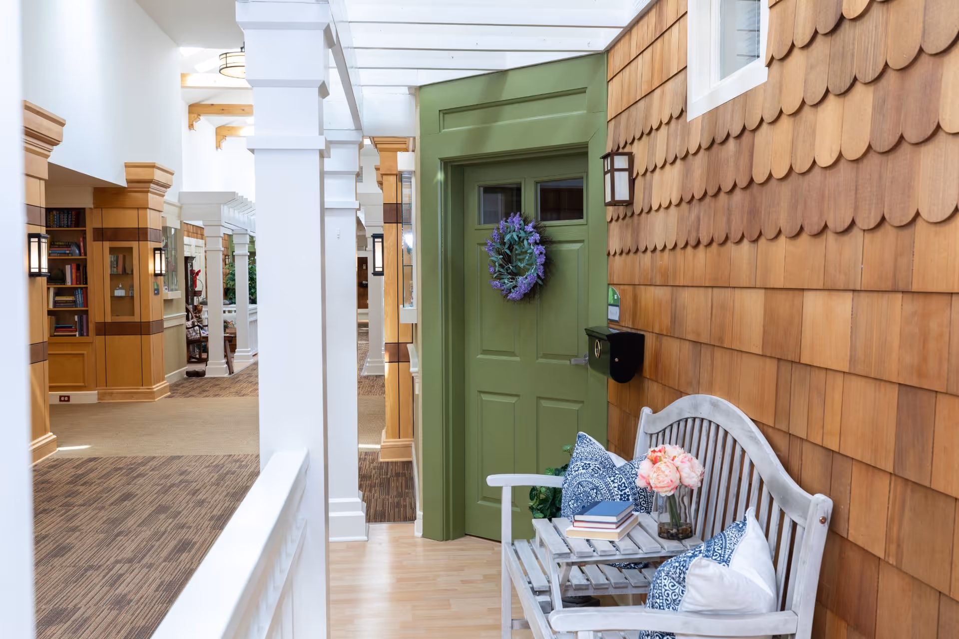 Indoor hallway area with a green door decorated with a purple wreath. Next to the door is a wooden bench with blue and white patterned pillows, a small table with books, and a vase of pink flowers. The walls have wooden shingles and the hallway features white columns and built-in wooden shelves with books and decor.