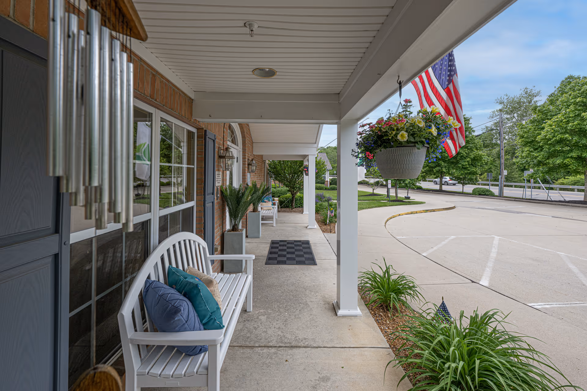Covered porch area with white benches adorned with colorful pillows, potted plants, hanging flower baskets, an American flag, and a wind chime. The porch overlooks a parking lot and landscaped greenery under a partly cloudy sky.