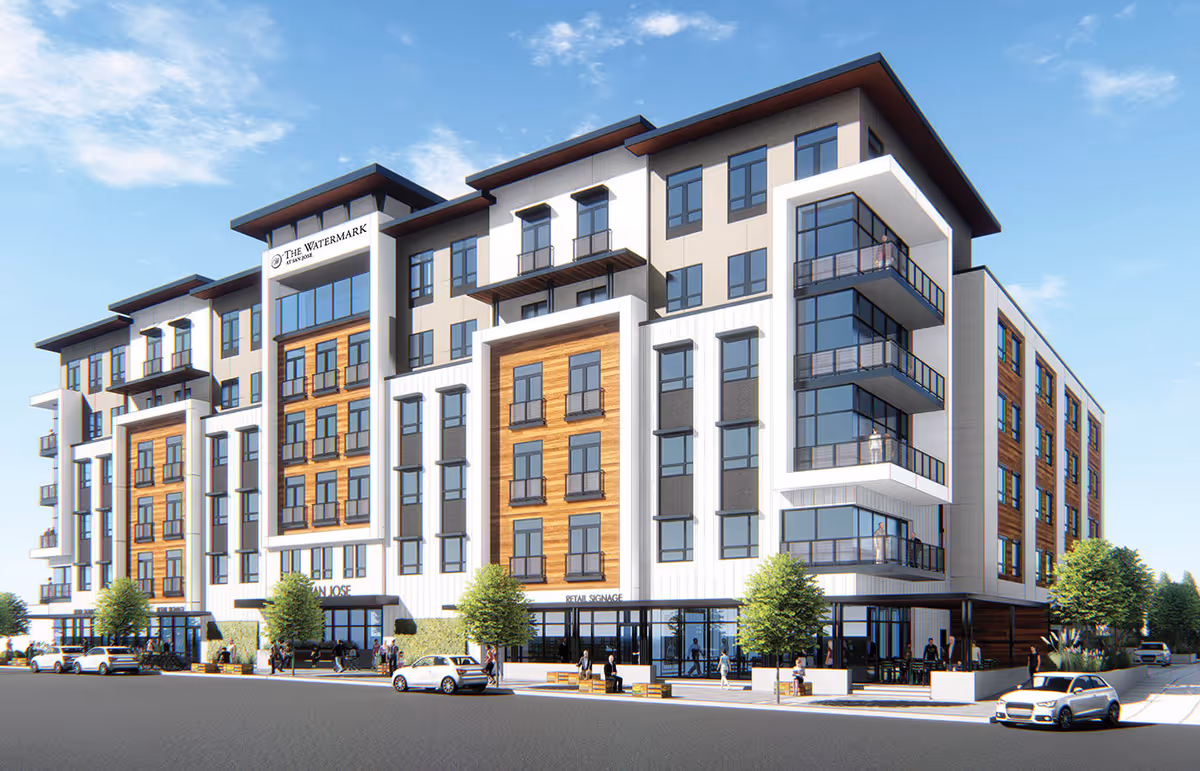 Modern multi-story senior living facility building with large windows, balconies, and retail spaces on the ground floor. Several people and cars are visible outside under a clear blue sky.