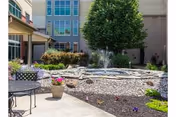 Outdoor courtyard area at a senior living facility with a small fountain surrounded by rocks and plants, a tree, and patio furniture including a table and chairs. The building with multiple windows is visible in the background.