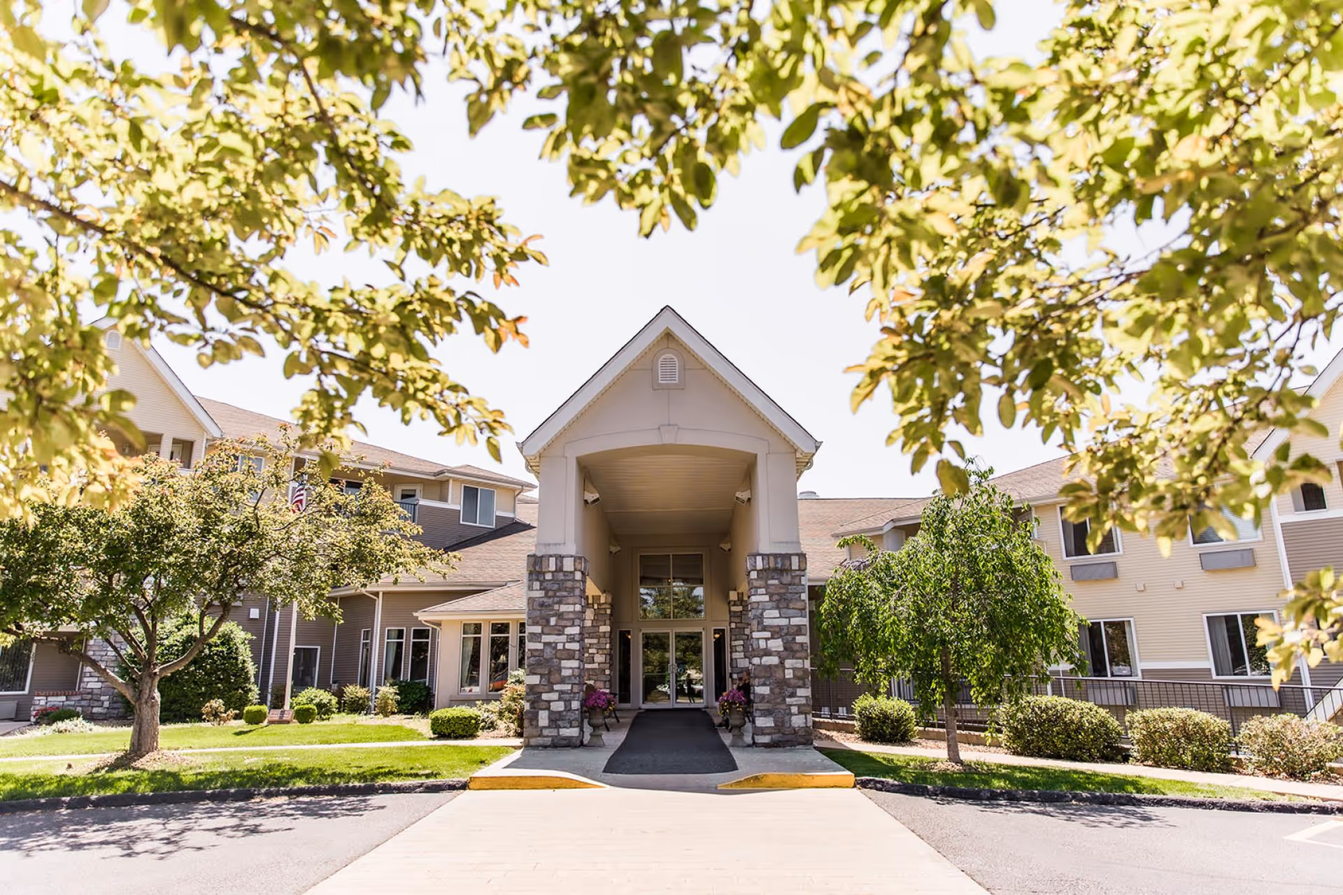 Front exterior view of College View Manor building with a covered entrance supported by stone pillars, surrounded by green trees and shrubs under a clear sky.