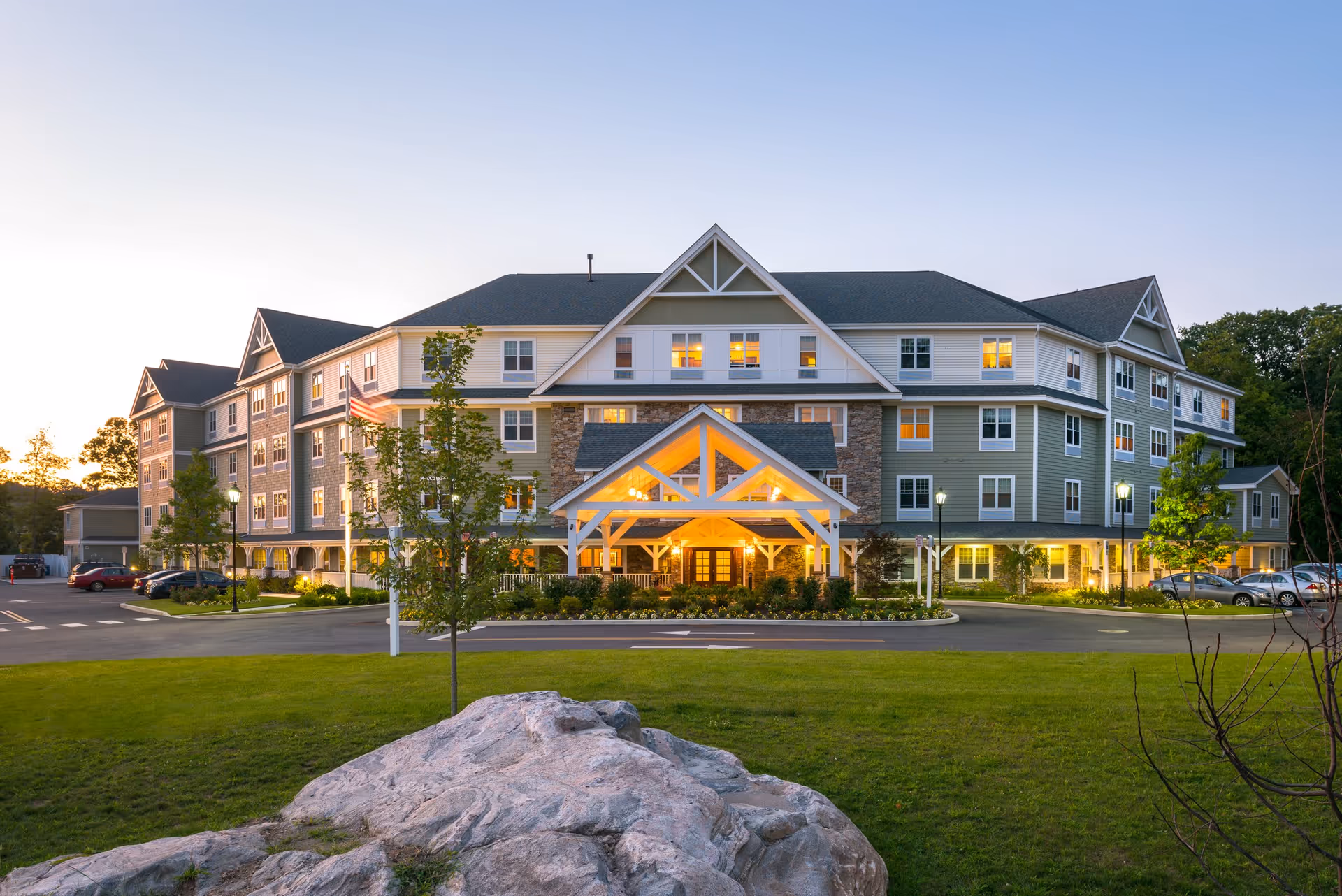 Exterior view of a large senior living facility building at dusk with lights glowing warmly from the windows and entrance. The building has a pitched roof, multiple windows, and a covered entrance with white beams. There is a well-maintained lawn with a large rock and some trees in the foreground, and a parking area with cars on either side of the building.