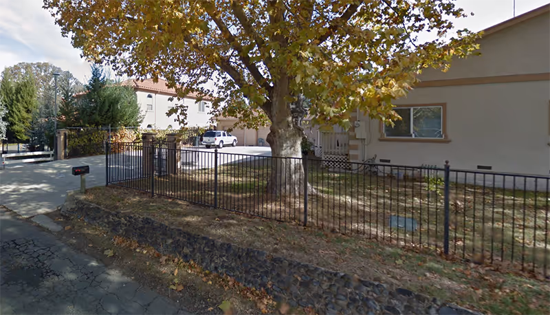 Exterior view of Golden Crest Care Center showing a fenced yard with a large tree, a stone retaining wall, a driveway, and a beige building with windows in the background.