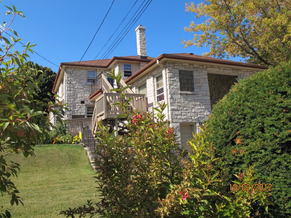 Exterior view of a two-story stone building with a brown roof, surrounded by green bushes and trees under a clear blue sky. There is a wooden staircase leading to the upper floor and a grassy lawn in the foreground.