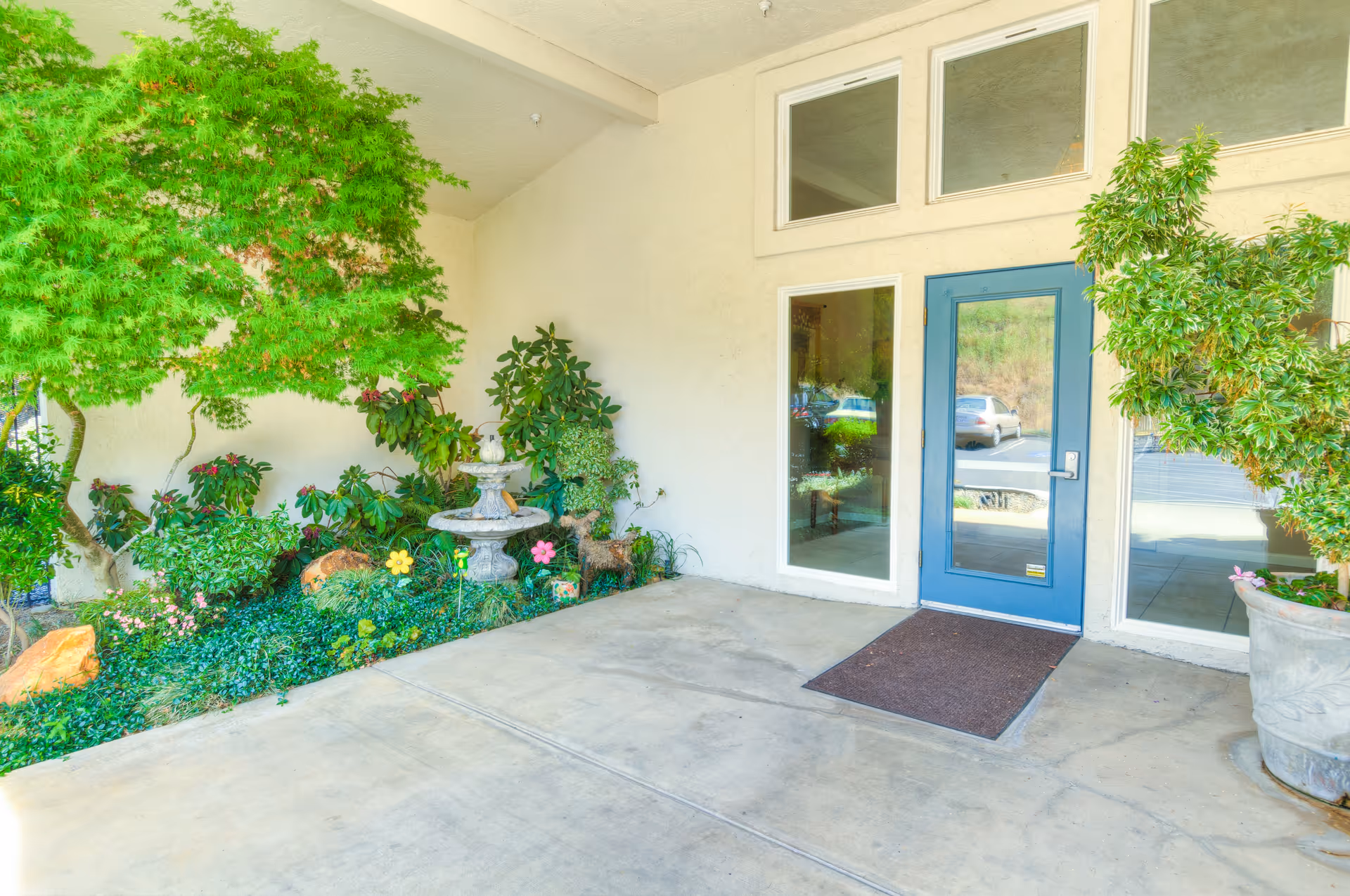 Entrance area of Oak Terrace Memory Care featuring a blue door with glass panels, surrounded by windows. There is a concrete floor with a brown doormat, and lush green plants and shrubs on either side, including a small stone fountain on the left side.
