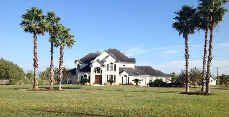 Large two-story white house with a dark roof set in a spacious green lawn with several tall palm trees under a clear blue sky.