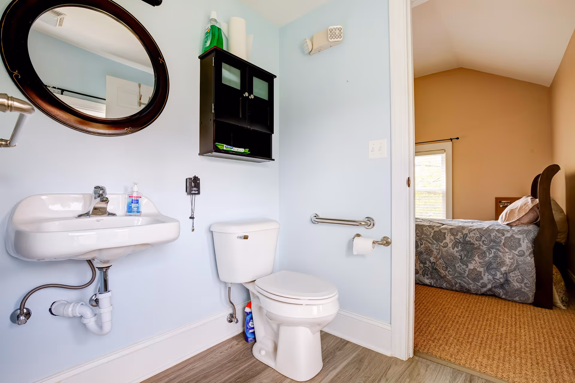A clean bathroom with a white sink and toilet, a black wall-mounted cabinet above the toilet, a round mirror above the sink, and a doorway leading to a bedroom with a bed covered in a patterned bedspread.