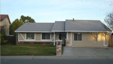 Single-story residential building with a gray roof, beige exterior walls, and a front yard with grass. There is a driveway leading to a garage on the right side and a mailbox on a brick post near the street.
