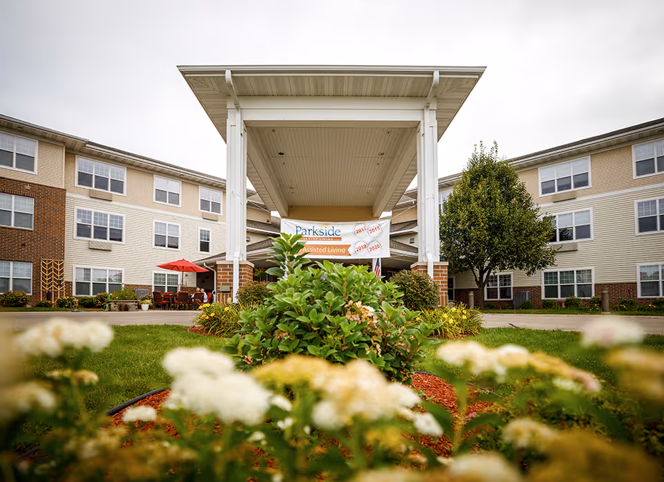 Front entrance of Parkside Senior Living facility with a covered drop-off area, surrounded by green bushes and flowers in the foreground, and a three-story building with multiple windows in the background.