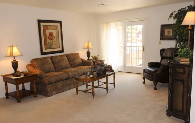 A cozy living room with a floral-patterned sofa, two wooden side tables with lamps, a wooden coffee table with decorative items, a dark leather armchair, a tall plant, framed artwork on the walls, and a glass door with sheer curtains leading to a balcony.