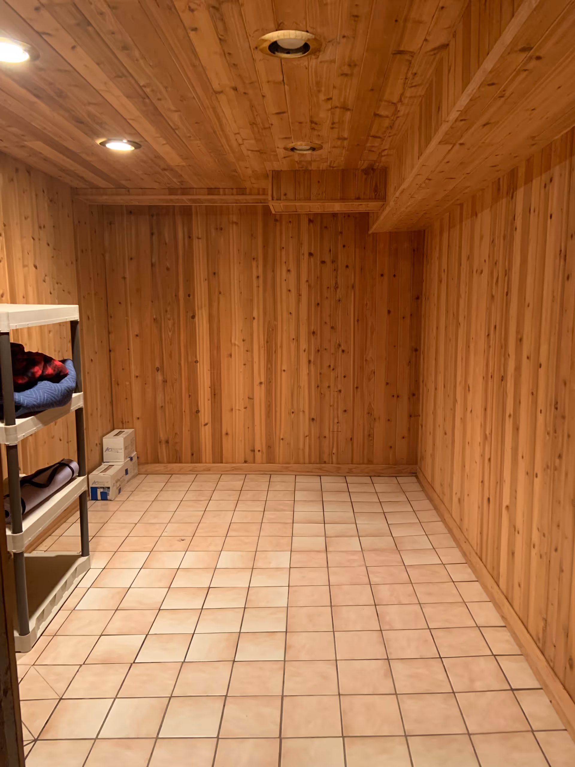 A small room with wood-paneled walls and ceiling, tiled floor, recessed ceiling lights, and a plastic shelving unit holding folded blankets and boxes in the corner.
