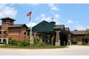 Exterior view of Valley Hi Nursing & Rehabilitation for McHenry County building with a covered entrance, brick walls, and an American flag on a flagpole in front under a partly cloudy sky.