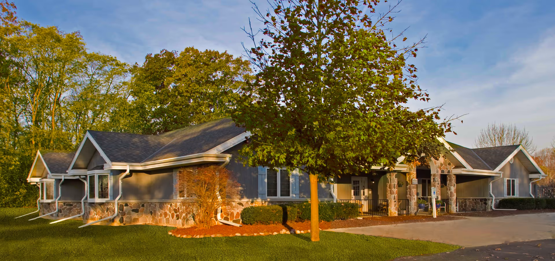 Exterior view of a single-story senior living facility building with stone and gray siding, surrounded by green trees and a well-maintained lawn under a blue sky.