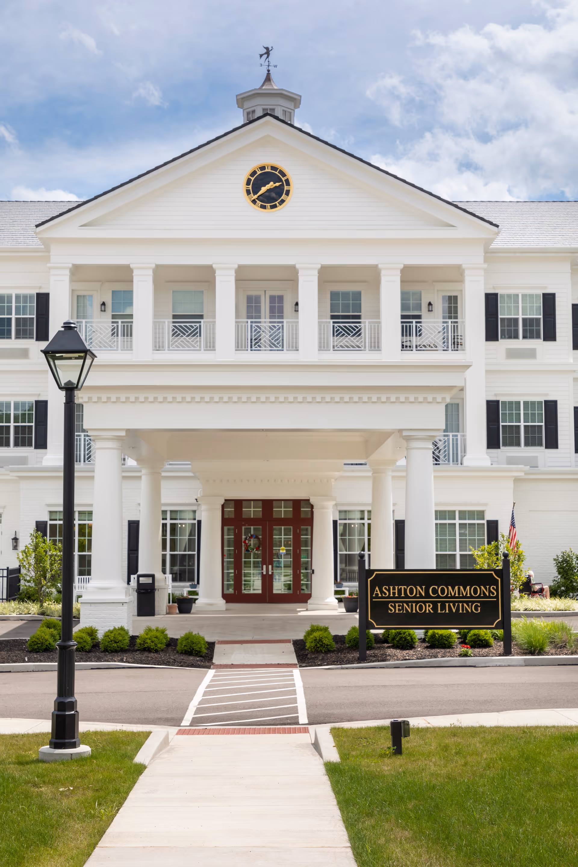 Front exterior view of Ashton Commons Senior Living building with white columns, a clock on the pediment, and a sign in front that reads 'Ashton Commons Senior Living'. The entrance has double doors and a covered driveway, with a lamppost and landscaped greenery nearby.