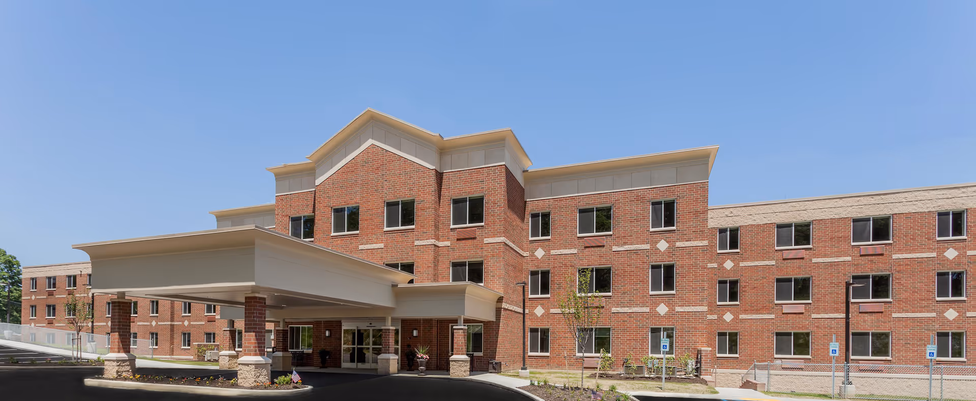 Front exterior view of a three-story senior living facility building with red brick walls, multiple windows, and a covered entrance driveway under a clear blue sky.