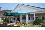Exterior view of San Simeon By The Sound Center For Nursing & Rehab building with a green awning over the entrance, white columns, and surrounding bushes and plants.