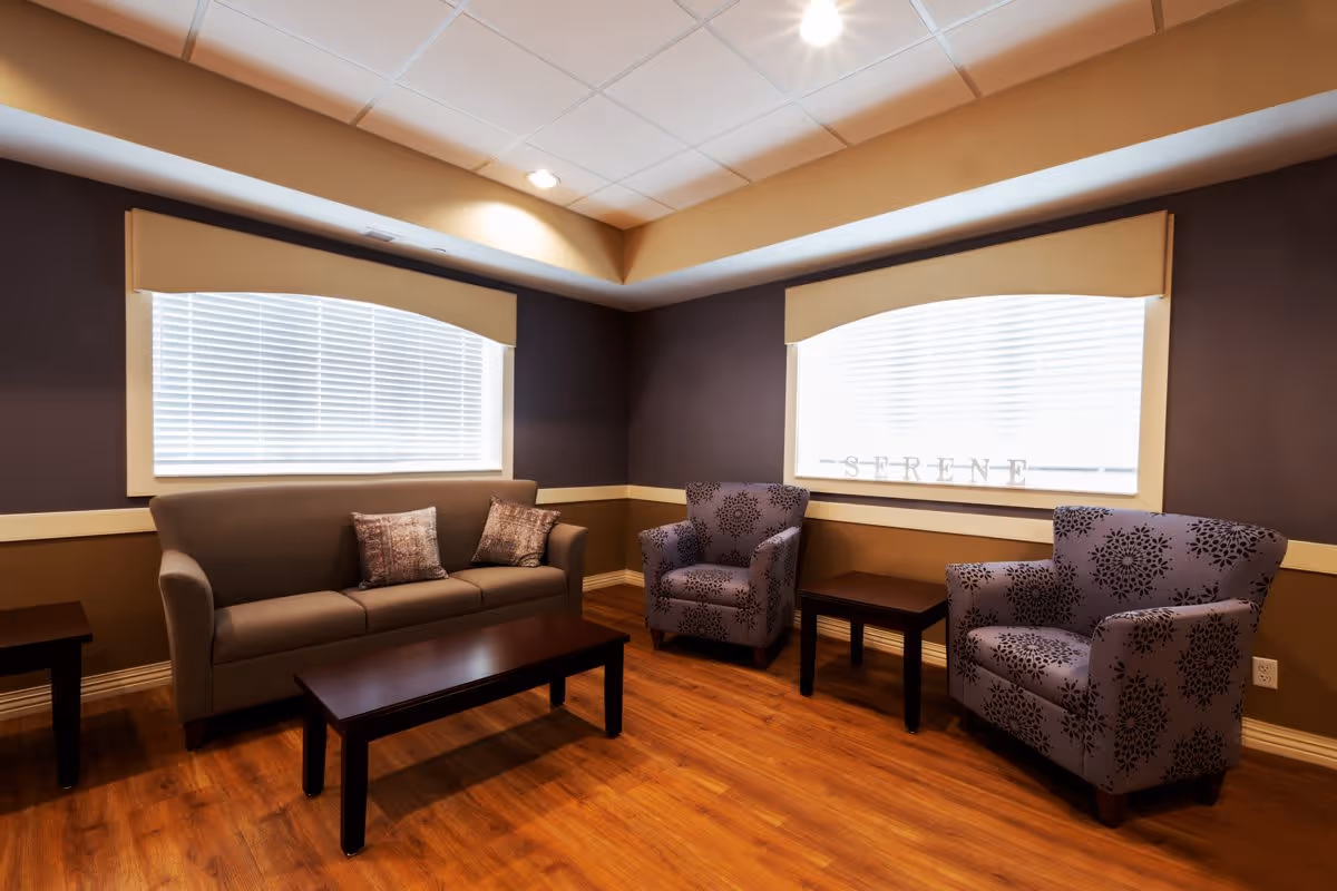 A cozy living room area with a brown sofa adorned with two patterned cushions, two purple armchairs with floral designs, a wooden coffee table, and two side tables. The room has wooden flooring, beige and dark brown walls, and two large windows with white blinds and beige valances. The word 'SERENE' is displayed on the window sill.