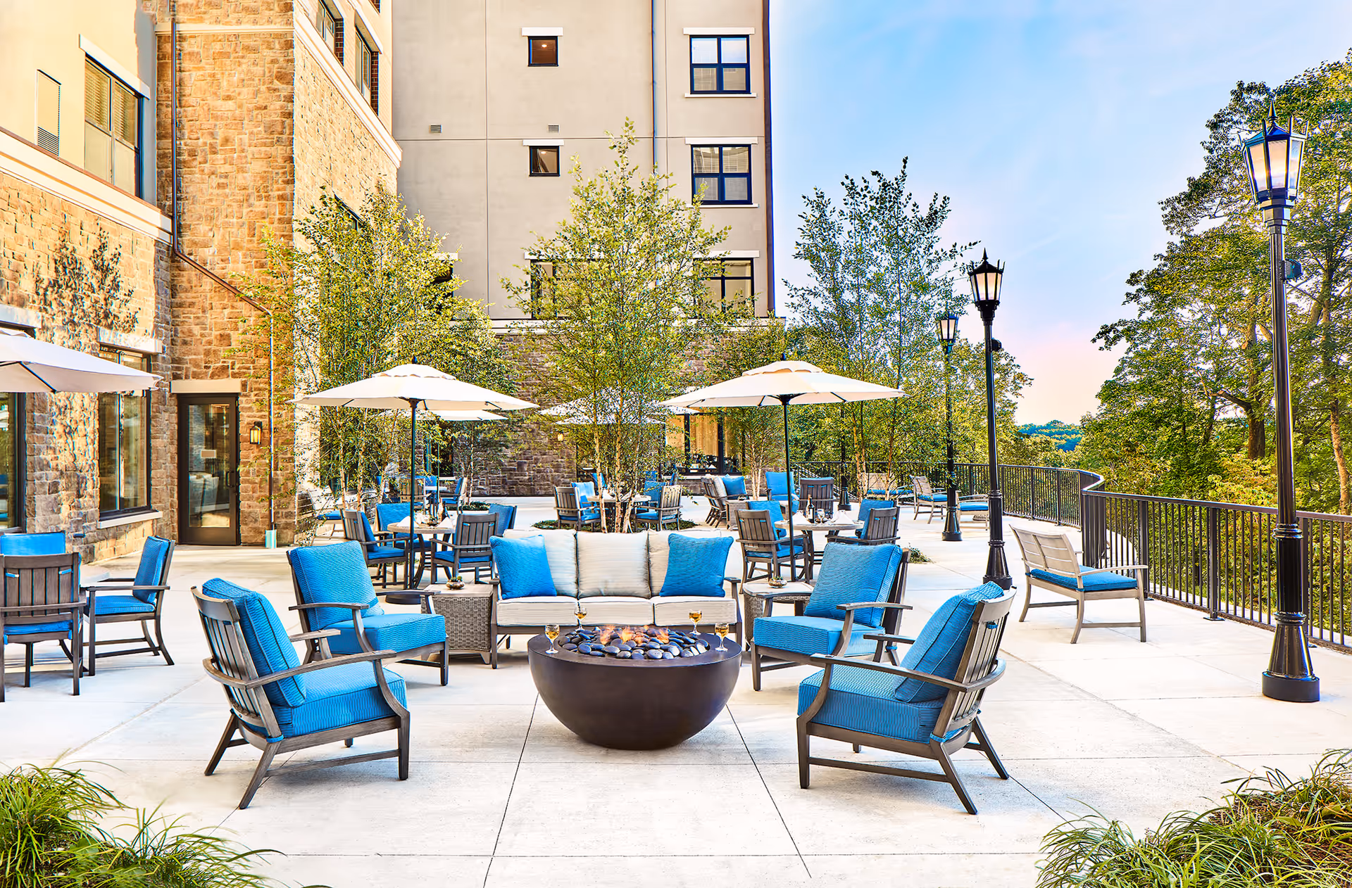 Outdoor patio area at The Club at Briarcliff Manor with blue cushioned chairs and sofas arranged around a fire pit. Several tables with umbrellas and chairs are set up along the patio. The area is surrounded by trees and lampposts, with a building wall on one side and a railing overlooking greenery on the other.