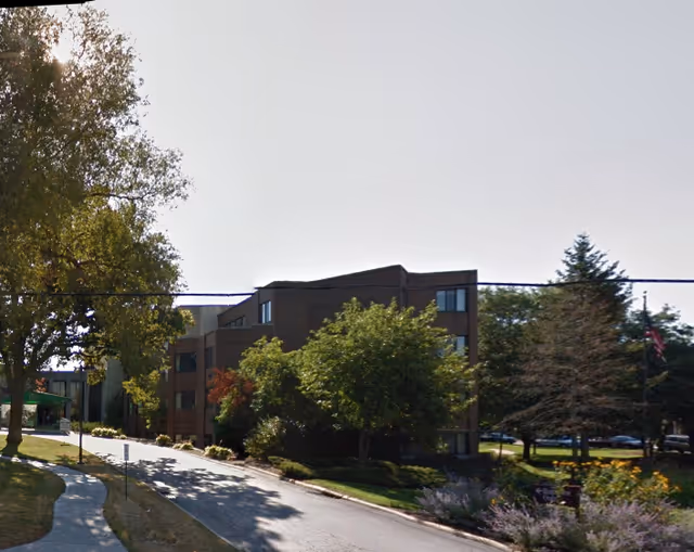 Exterior view of a multi-story senior living facility building surrounded by trees and landscaping, with a driveway leading up to the entrance.