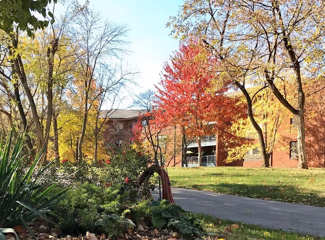 Outdoor view of a senior living facility named Our Lady of Life Apartments showing a paved pathway surrounded by green grass, various trees with autumn foliage in shades of yellow, orange, and red, and a brick building in the background under a clear blue sky.