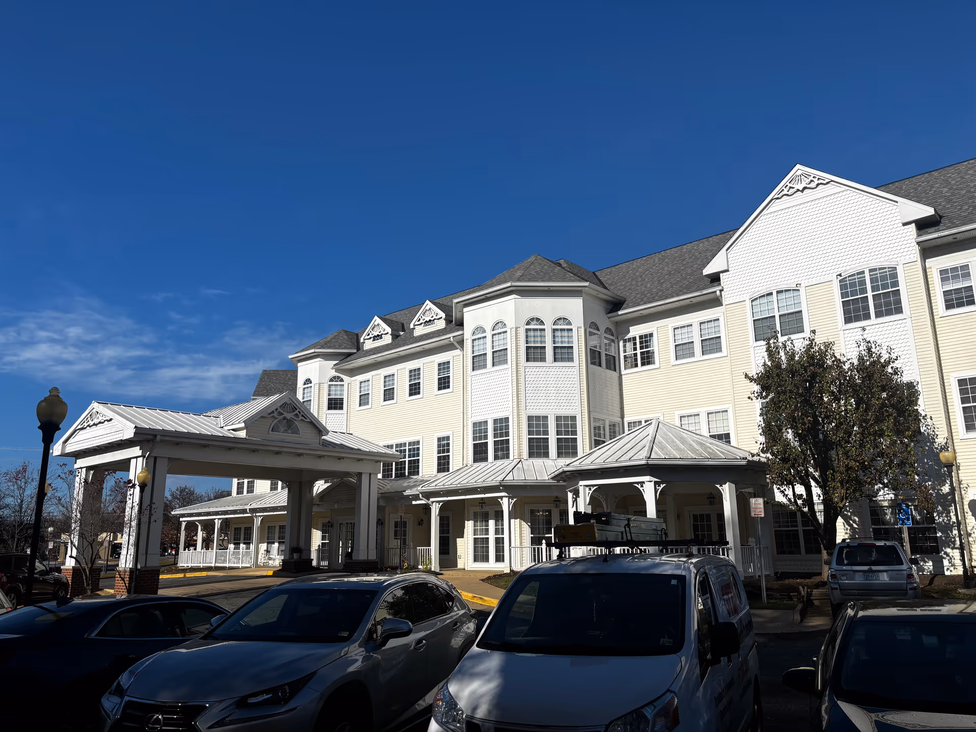 Exterior view of a large, multi-story senior living facility building with white siding and multiple windows under a clear blue sky. Several cars are parked in front of the building near a covered entrance.