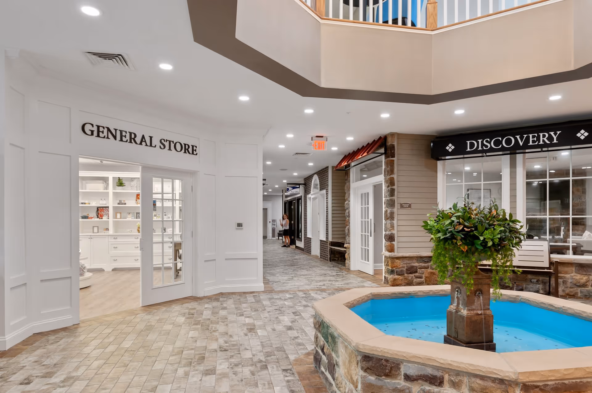 Interior view of a senior living facility hallway with a general store on the left and a discovery area on the right. The hallway features a stone-tiled floor, a central decorative fountain with greenery, and bright overhead lighting. A person is visible in the distance walking down the hallway.