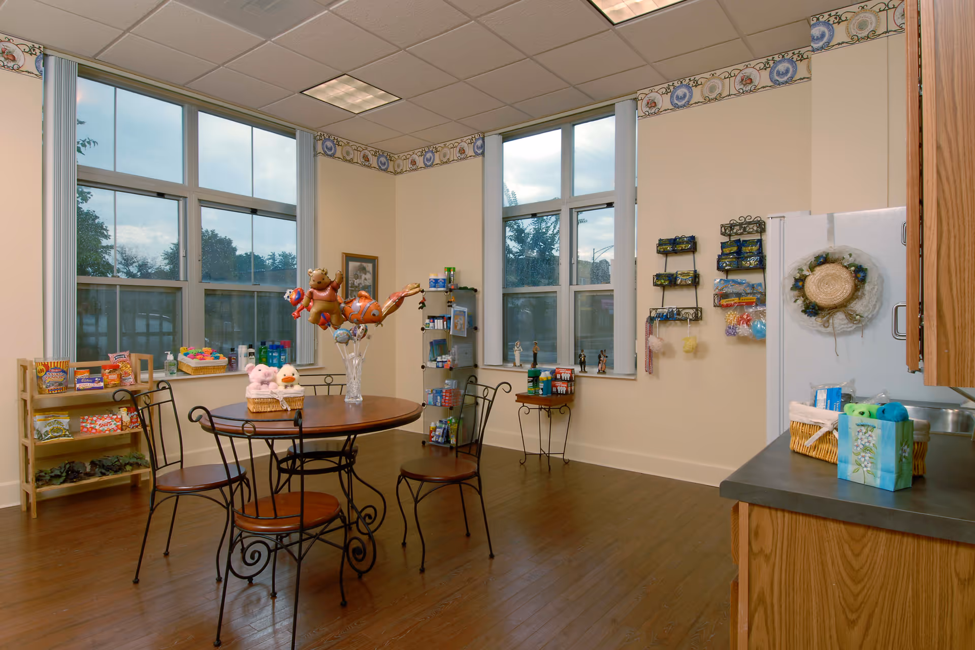 A cozy room with large windows letting in natural light, featuring a round wooden table with four metal chairs. On the table are a vase with animal-shaped balloons and a basket with plush toys. Shelves and racks along the walls hold various snacks, toiletries, and small items. The room has wooden flooring and light-colored walls with decorative border trim near the ceiling.