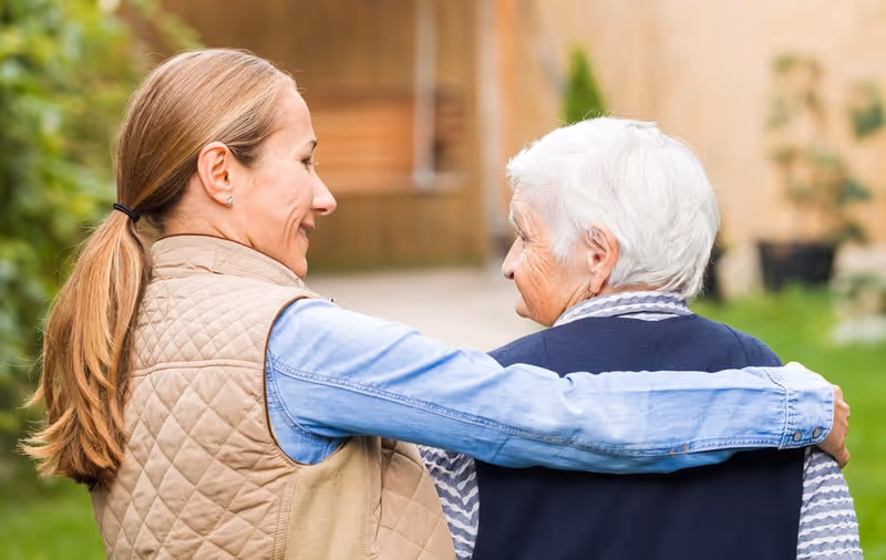 A younger woman with a ponytail and a beige quilted vest has her arm around an elderly woman with short white hair wearing a navy blue sweater. They are outdoors, walking together and looking at each other with smiles.