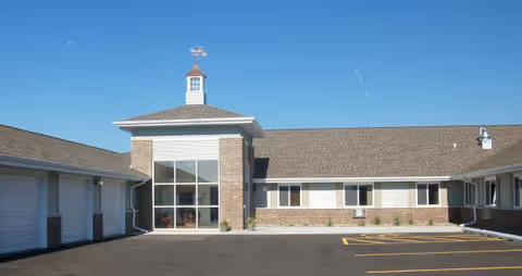 Exterior view of a single-story building with a central entrance featuring large windows and a small cupola on the roof, surrounded by a paved parking area under a clear blue sky.