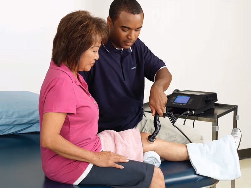 A healthcare professional is using a therapeutic ultrasound device on the knee of a seated woman in a clinical setting. The woman is wearing a pink shirt and dark pants, and the professional is focused on the treatment. The woman's leg is supported with towels and a medical device is placed on a nearby table.
