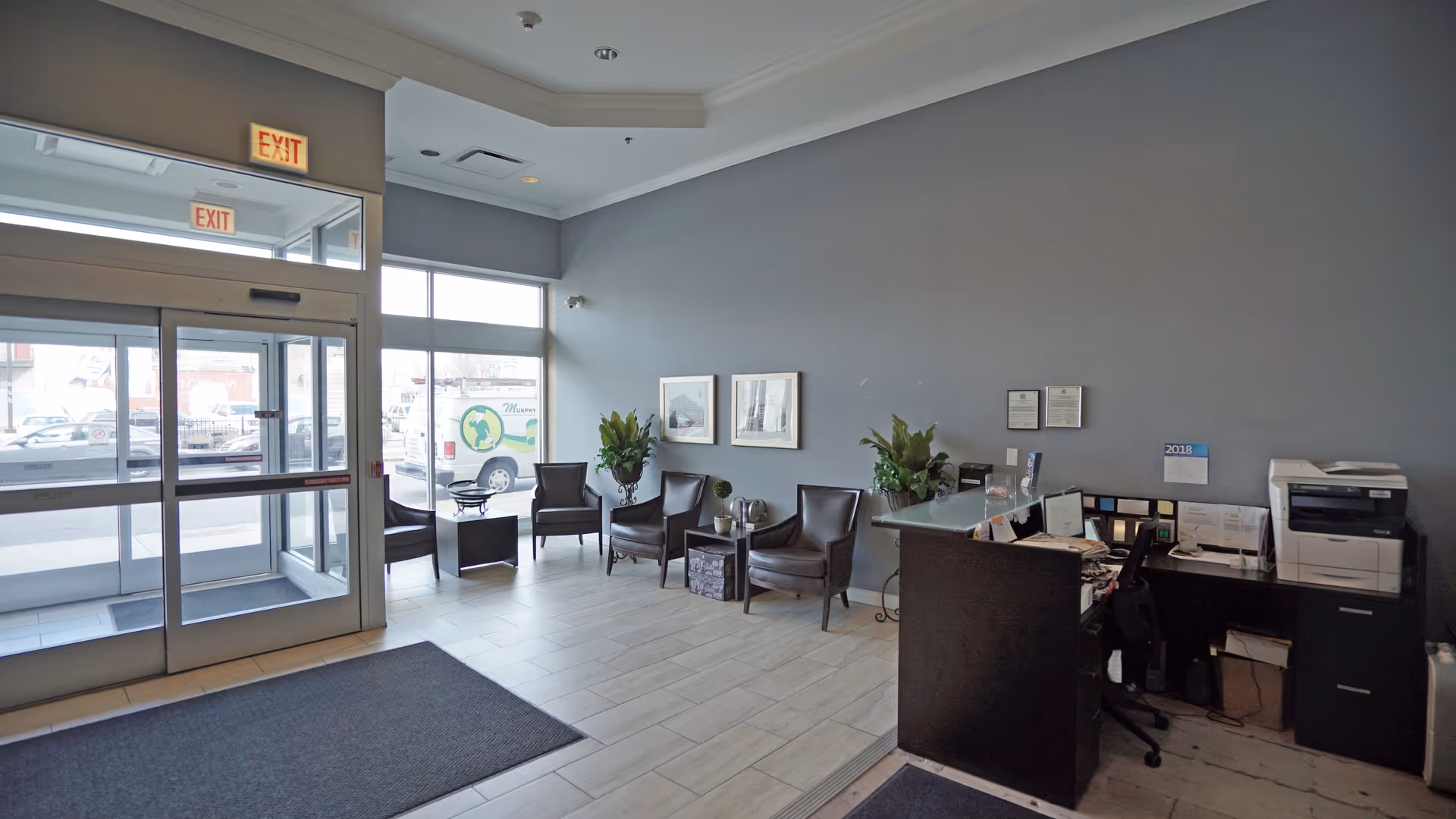 Reception area of a facility with a dark wooden desk, office chair, printer, and paperwork. There are four dark leather chairs arranged along the wall with two framed pictures above them and two potted plants. Large windows and glass doors provide natural light and a view of parked cars outside. Exit signs are visible above the doors.