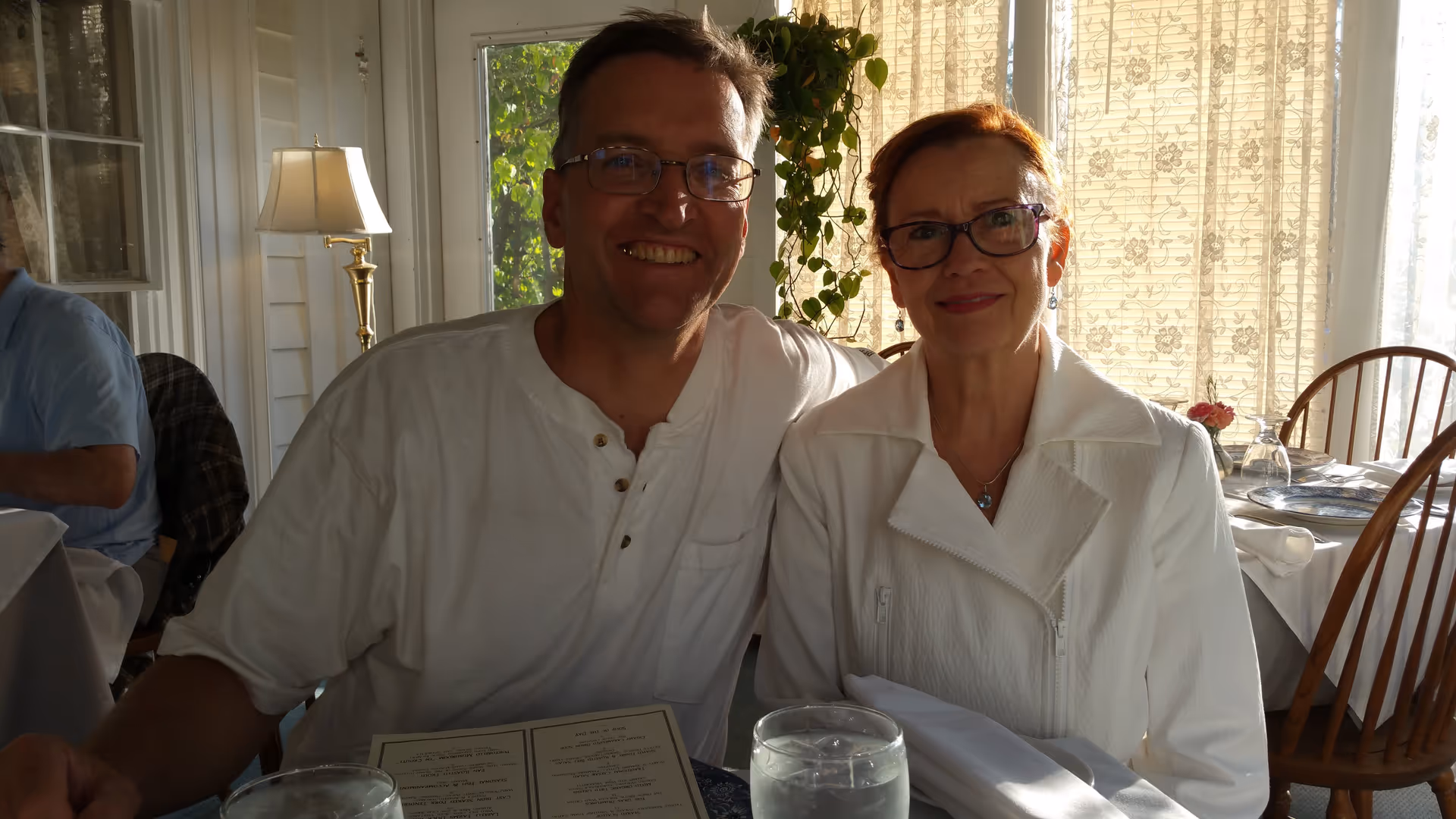 A man and a woman sitting closely together at a dining table in a well-lit room with large windows and floral curtains. The man is wearing glasses and a white shirt, smiling broadly, while the woman is also wearing glasses and a white jacket, smiling gently. There are wooden chairs and a table set with plates, glasses, and napkins in the background.