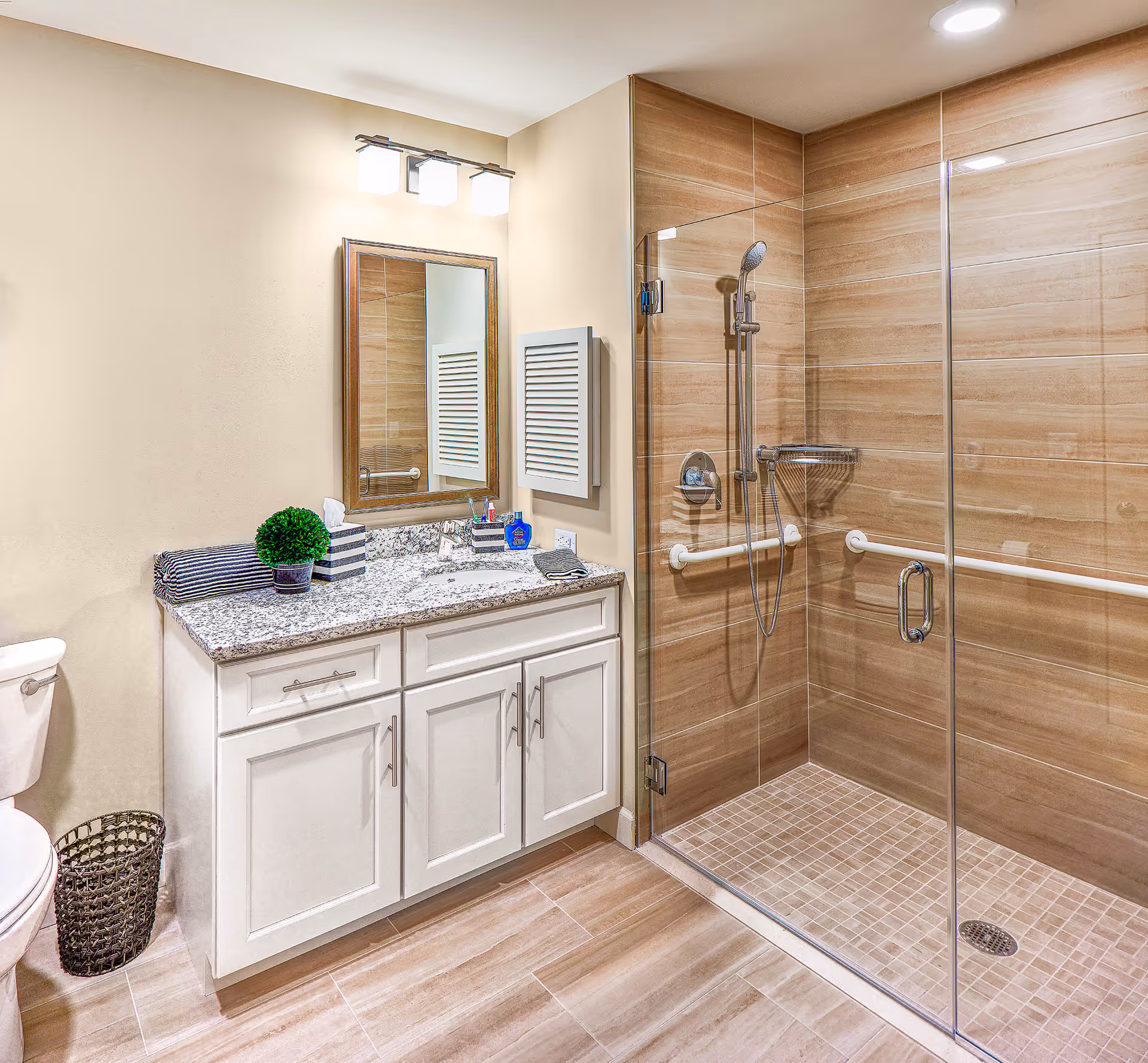 A modern bathroom featuring a glass-enclosed shower with beige tiles and grab bars, a white vanity with granite countertop, a mirror with three light fixtures above it, a toilet, and a small waste basket on the floor.