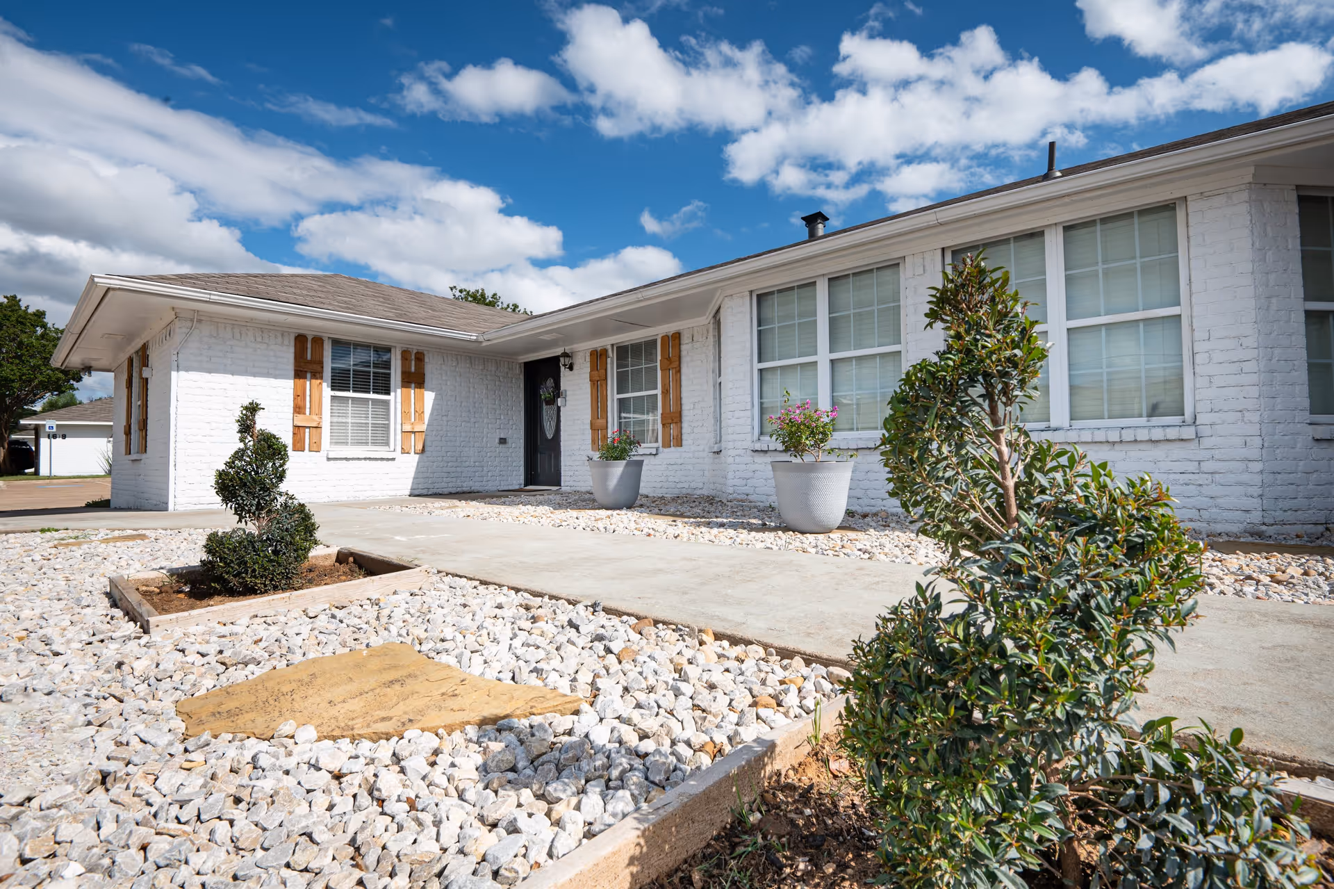 Exterior view of a single-story white brick building with wooden shutters on the windows, a black front door, and a landscaped front yard featuring white rocks, small shaped bushes, and potted plants under a partly cloudy blue sky.